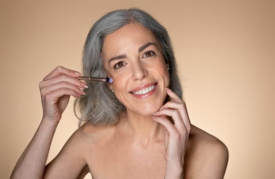 Beautiful senior caucasian woman holding eye shadow brush, doing daily makeup, smiling at camera over beige background