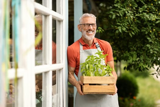 Portrait of handsome mature male gardener carrying plants in crate outside greenhouse