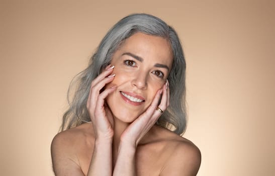 Senior grey-haired woman touching her face and smiling at camera, doing morning face care routine, beige background