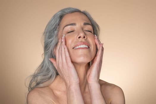 Spa treatments. Good-looking senior woman with beautiful skin touching her face, enjoying beauty care while posing over beige studio background