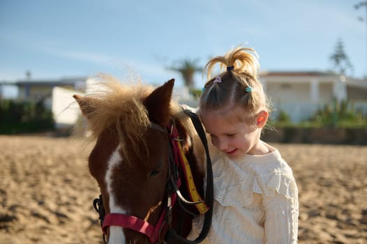 Cute blonde little child girl stroking a pony, smiling and enjoying happy weekend outdoor. Children and love for animals