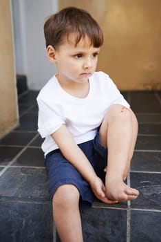 Little boy, sitting and injury on knee in home and accident, ache and with blood from cut. Young child, sad and holding hurt leg up with hand on foot, pain and sore on bruise with fear of trouble
