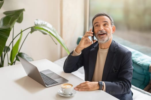 Cheerful senior businessman on phone with laptop and coffee