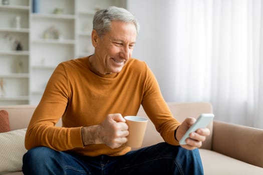 Smiling old man sitting on couch at home, holding phone