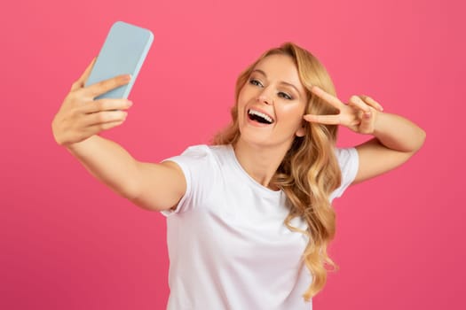 blonde woman making selfie on smartphone gesturing victory sign, studio