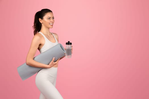 A smiling woman in a white tank top and leggings carrying a yoga mat and water bottle