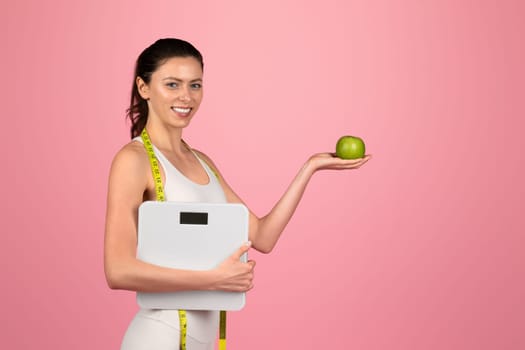 Health-conscious woman with a measuring tape around her neck holding a green apple and a scale