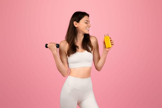 Happy athletic woman in white workout gear lifting a dumbbell and holding a bottle of orange juice