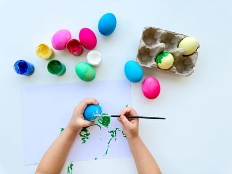Childrens hands paint Easter eggs with brush surrounded by colorful eggs and jars of paints on white table, creative holiday activity.