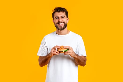 Happy European man posing holding a cheeseburger on yellow backdrop