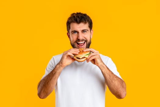 man biting delicious takeaway cheeseburger smiling to camera, yellow backdrop