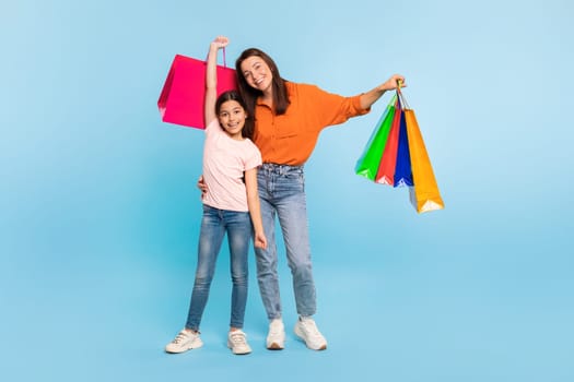 Happy young mother and daughter posing with colorful shopping bags standing together, enjoying successful shopping day, carrying purchases on blue backdrop, full length. Seasonal sales