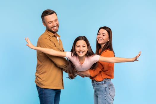 Happy family of three posing lifting kid daughter simulating flight, girl spreading her arms wide like plane, evoking joys of travel and vacation, having fun on blue background, studio shot