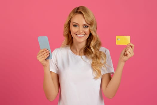 Happy blonde woman holding smartphone and credit card, pink background