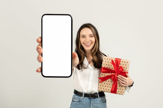 Joyful woman presenting a smartphone with a blank screen in one hand and holding a gift
