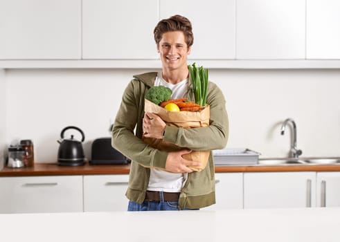Portrait, vegetables and man with brown paper bag, home and wellness with nutrition and vegetarian. Face, person and guy with groceries and healthy ingredients with products, food and diet in kitchen