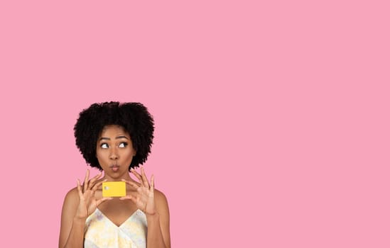 Amused African American woman with curly hair playfully holding a credit card to her lips