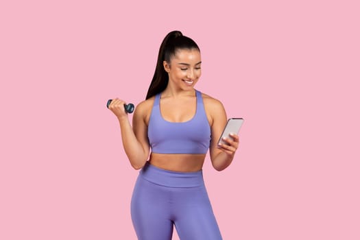 Smiling woman with dumbbell checking phone on pink background