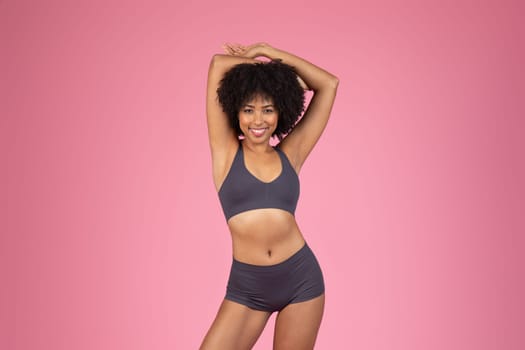Joyful black woman in fitness attire stretching on pink background