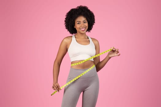 Joyful black woman measuring waist with tape on pink backdrop