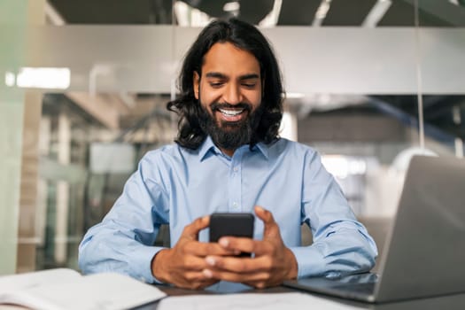 Positive indian guy manager using smartphone, modern office interior
