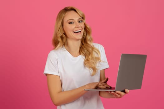 Happy blonde woman holding laptop over pink studio background