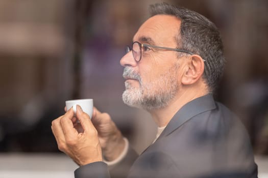 Contemplative mature man with glasses looking out the window, holding a small espresso