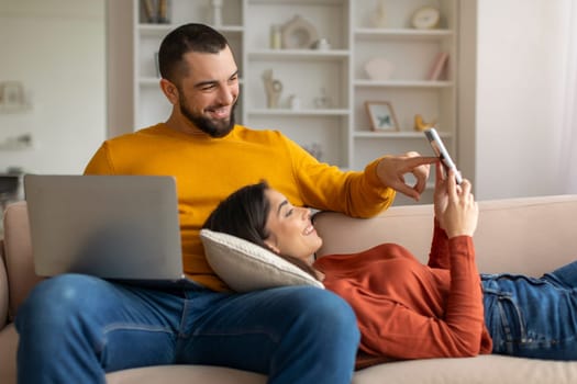 Happy Young Couple Reading Message On Smartphone And Using Laptop At Home