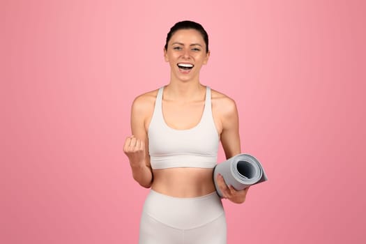 Exuberant young woman in white yoga attire holding a rolled-up yoga mat, ready for a workout session