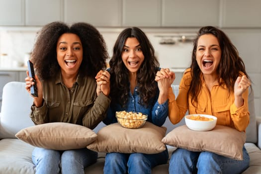 Three emotional diverse women cheerfully shouting reacting watching TV indoor