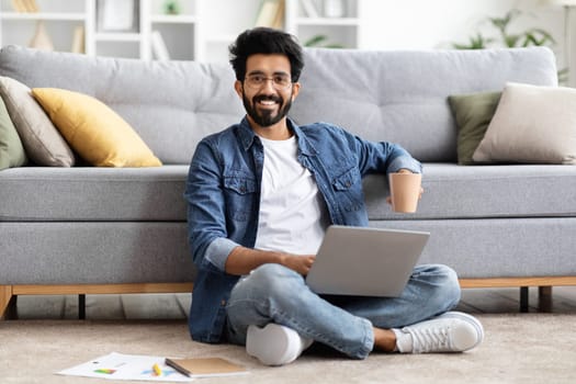 Relaxed indian man sipping coffee while working on laptop at home