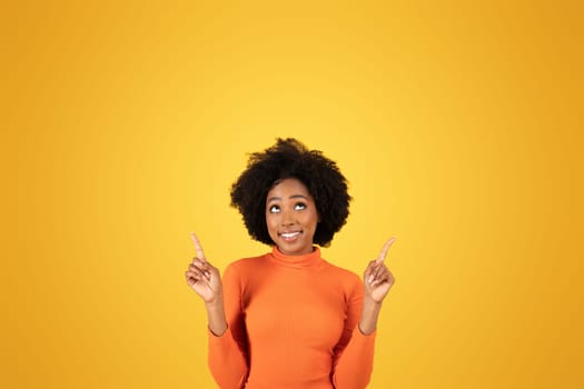Optimistic African American woman with afro hair looking up and pointing upwards, expressing positivity