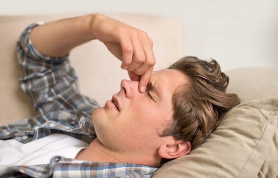 Man, nose and headache pain on sofa or sinus problem with fatigue or allergy infection, sick or bacteria. Male person, unhappy and tired migraine in London home or flu disease or stress, bug or dizzy