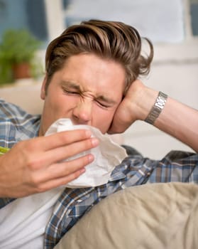 Sick, flu or man sneezing into tissue on sofa with cold, virus or hay fever at home. Influenza, allergies or person with toilet paper for viral infection, bacteria or coughing with sinusitis in house