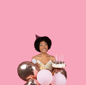 Joyful young woman with natural curly hair, wearing a birthday hat, holding balloons