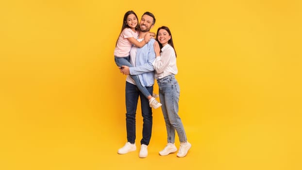 Family Of Three Posing On Yellow Backdrop, Father Carrying Daughter