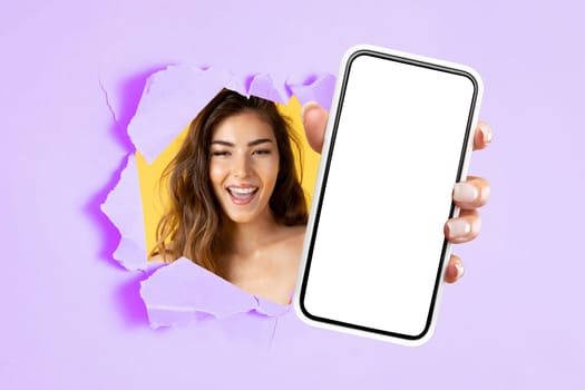 Radiant young woman with a beaming smile peeking through a purple and yellow torn paper, holding a smartphone