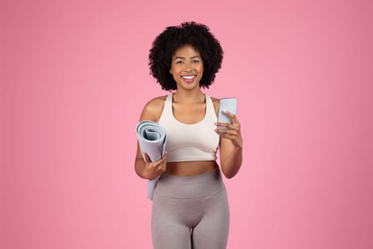 Cheerful black woman ready for workout, holding yoga mat under one arm and smartphone in the other hand, against vibrant pink background, embodying an active lifestyle