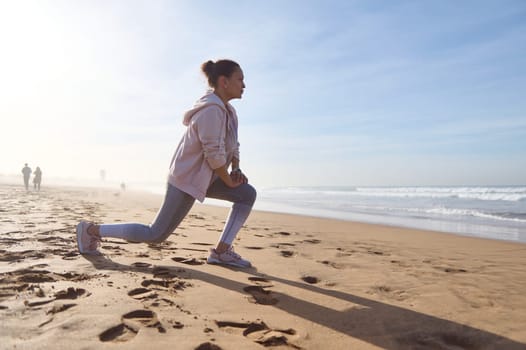 Active fitness woman doing lunges and warming her leg muscles while exercising on the sandy beach during morning workout