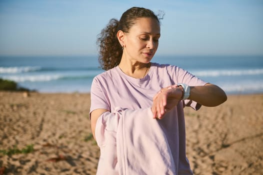 Young female athlete checking her heart rate with smartwatch, relaxing after running or bodyweight training on the beach