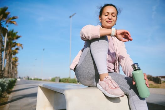 Fit woman checking digital watch for heart rate, pulse or cardio performance in nature, sitting on a stone bench outdoor