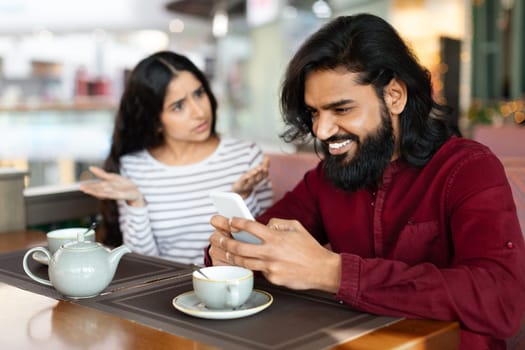 Angry young indian woman looking at boyfriend stuck in smartphone,