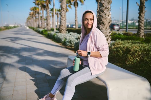 Attractive young woman sitting on a stone bench with bottle of water after outdoor workout and looking into the distance