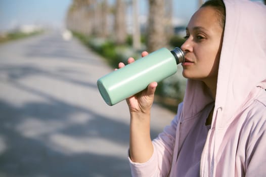 Close-up Latina sporty woman in purple hoodie, holding a steel bottle, drinking water to refresh her body after workout
