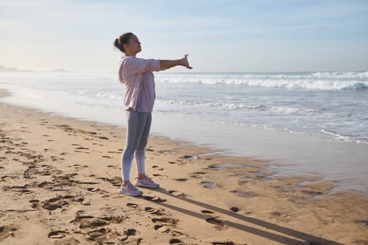 Full length sporty woman in activewear exercising on the beach, stretching and warming up her body before morning jog