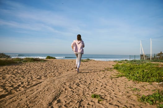 Back view of sporty woman jogging on sand towards ocean during morning run. Sport. Fitness. Active healthy lifestyle