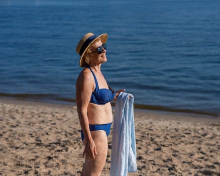 An old woman in a straw hat, sunglasses and a swimsuit is resting on the beach.
