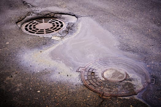 An oil slick against the background of an asphalt road flows into a storm drain against the background of a sewer grate and the lid of a closed well.
