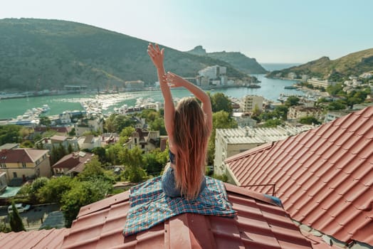 Woman sits on rooftop with outstretched arms, enjoys town view and sea mountains. Peaceful rooftop relaxation. Below her, there is a town with several boats visible in the water