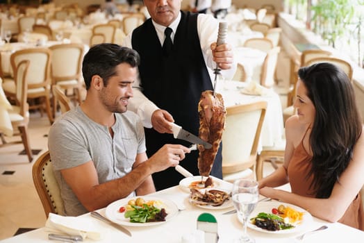 Happy couple, date and dining with kebab of waiter serving, meat or slices on romantic dinner at table. Young man or woman with chef, skewer or food for meal, eating or enjoying service at restaurant